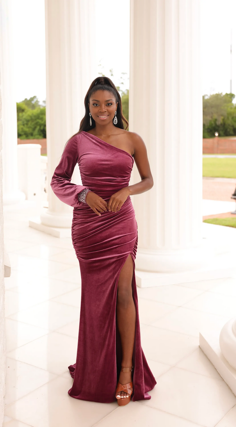 This dress features a one-shoulder neckline with a long sleeve embellished with a beaded wrist, a ruched torso for a flattering fit, velvet fabric, and a slit. This gown is elegant and timeless. Choose this stunning dress for your next prom or formal event.  STS 1023