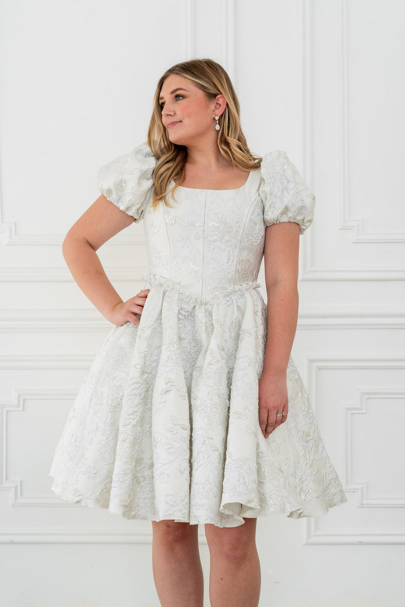This dress features a shimmering brocade fabric that catches the light with every movement. The hidden structured boning and soft square neck provides a flattering, corset-like fit, while the full zipper back ensures effortless wear. The puff sleeves add a touch of romance, making it perfect for your next homecoming or formal event.

AG 11003